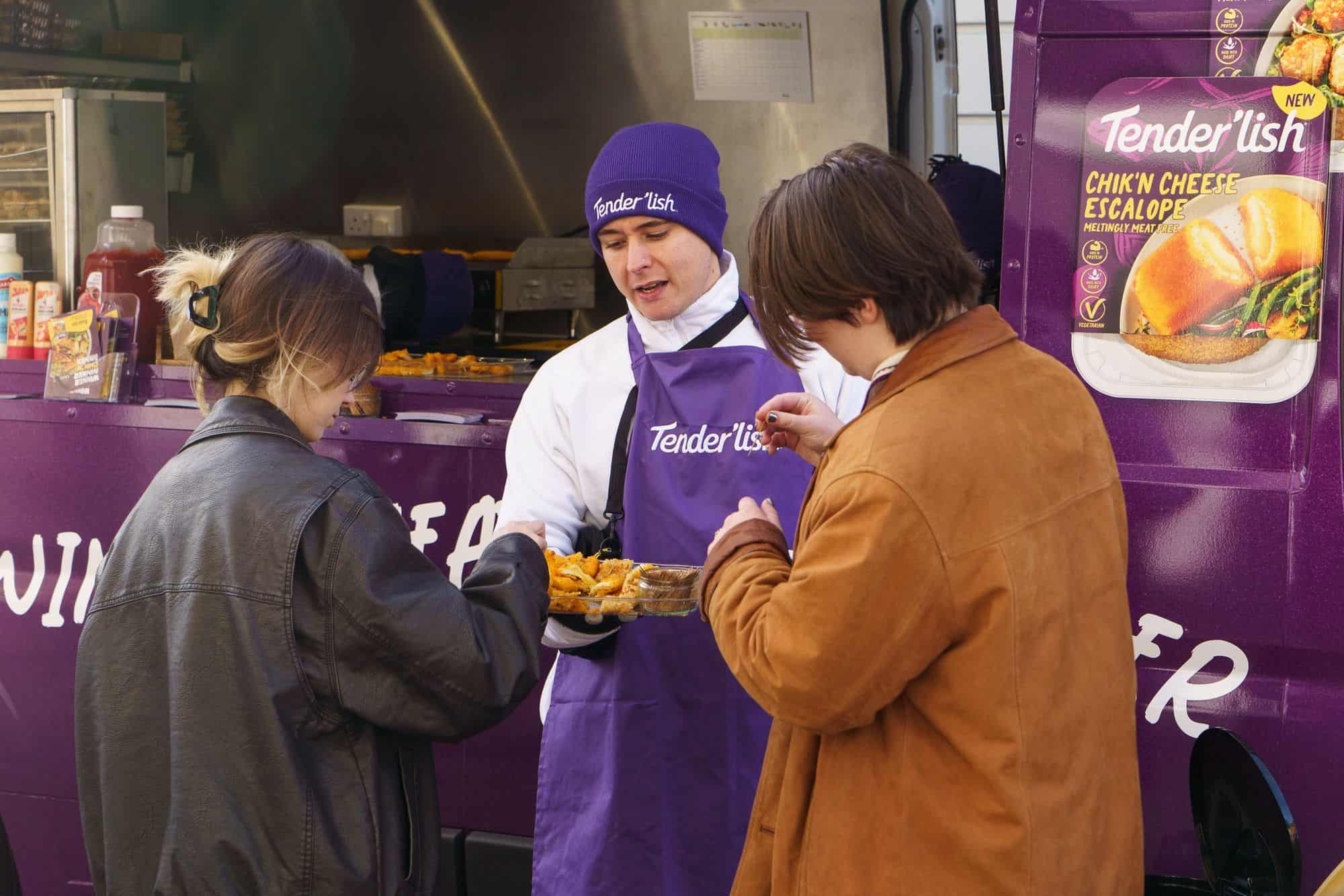 A brand ambassador serving hot meat-free samples from a purple Tender'lish food truck to two people
