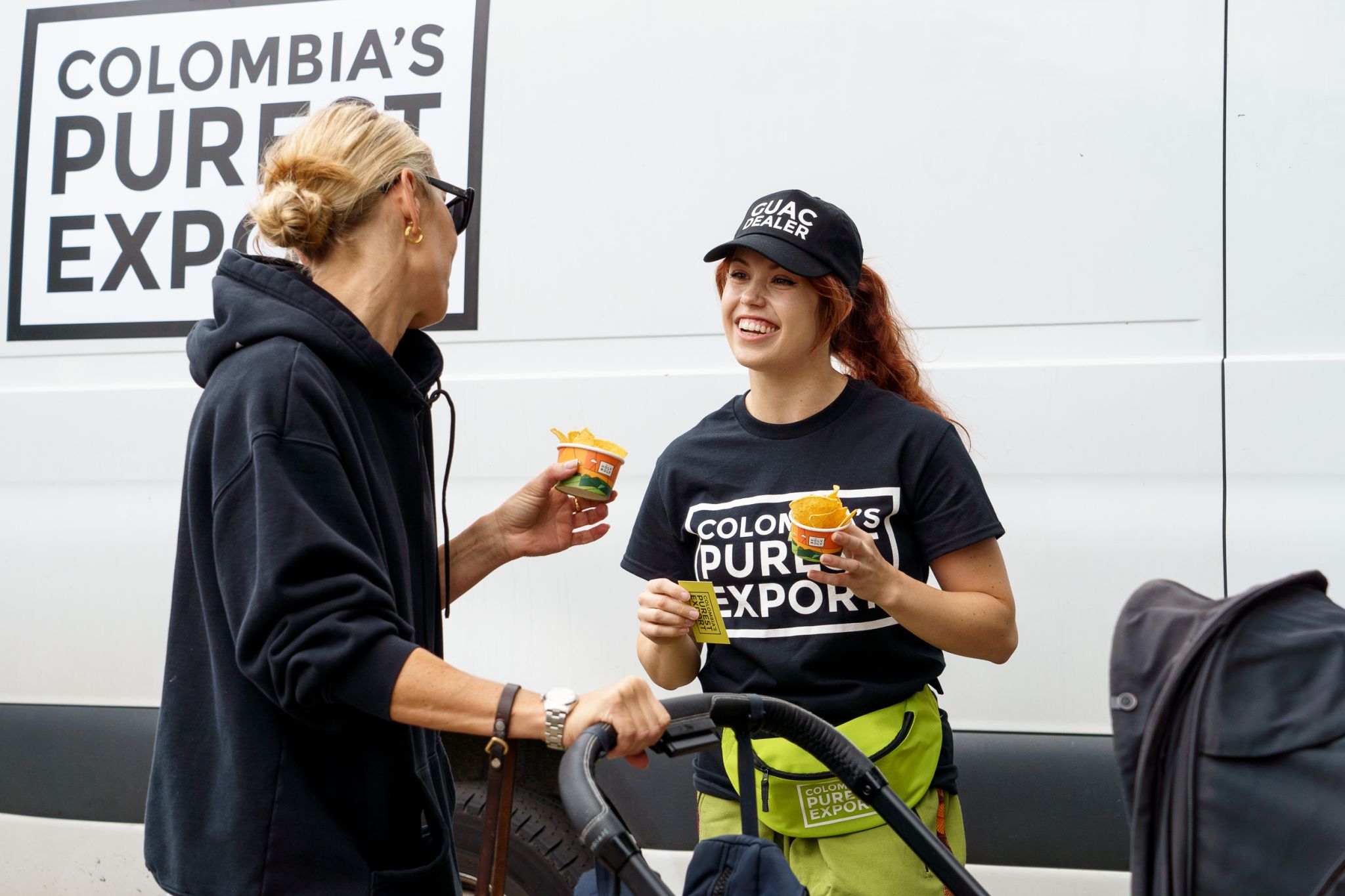 A Holy Moly brand ambassador offering guacamole samples to a woman with a stroller on a city street.